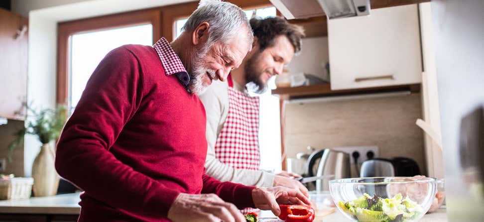 An older man and his son are cooking together in the kitchen, both smiling and enjoying each other's company. 