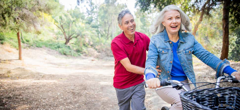 An older woman is being pushed by her husband while riding her bike outside in nature.