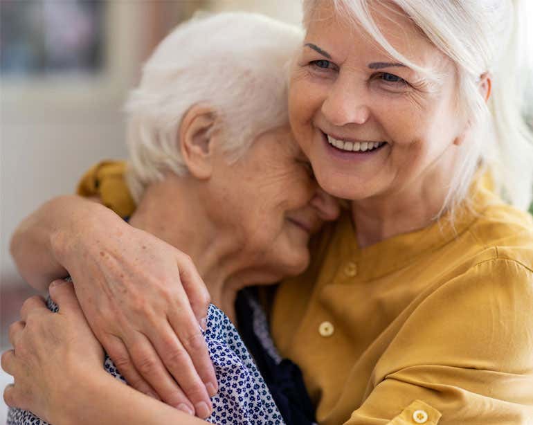 A Caucasian senior woman embraces her mother while smiling.