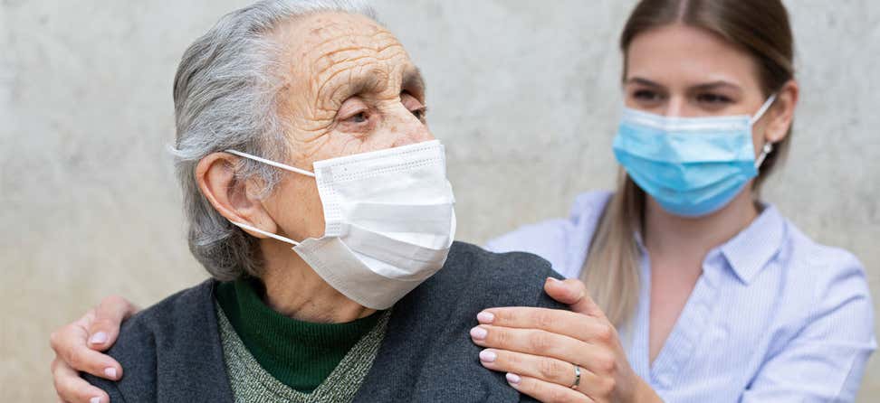 A younger female caregiver puts her hands on the shoulders of an elderly woman wearing a mask because of COVID-19 pandemic.