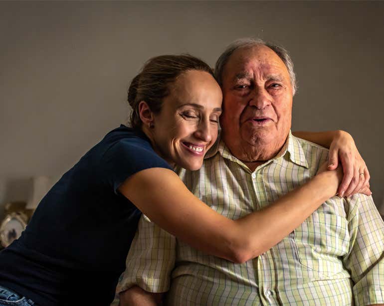 A Hispanic young, female caregiver hugs her senior father while sitting at home.