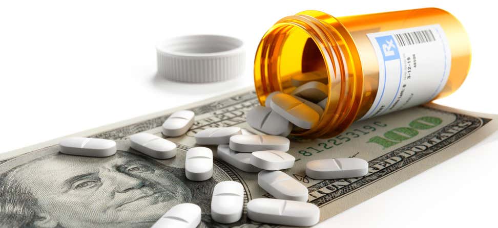 A close up shot of a prescription bottle and pills scattered atop a one-hundred dollar bill.