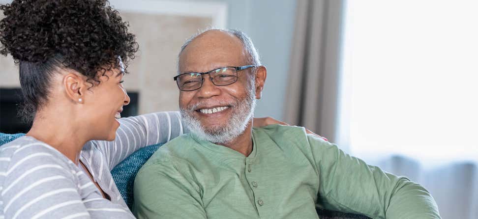 A senior Black man is smiling while looking at his middle-aged female caregiver.