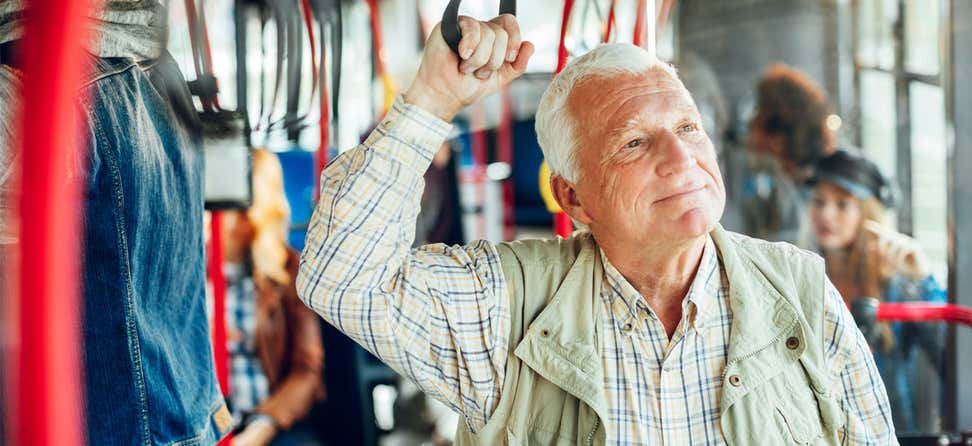 A senior man, wearing plaid and other outerwear, is standing up on a bus and enjoying his day.