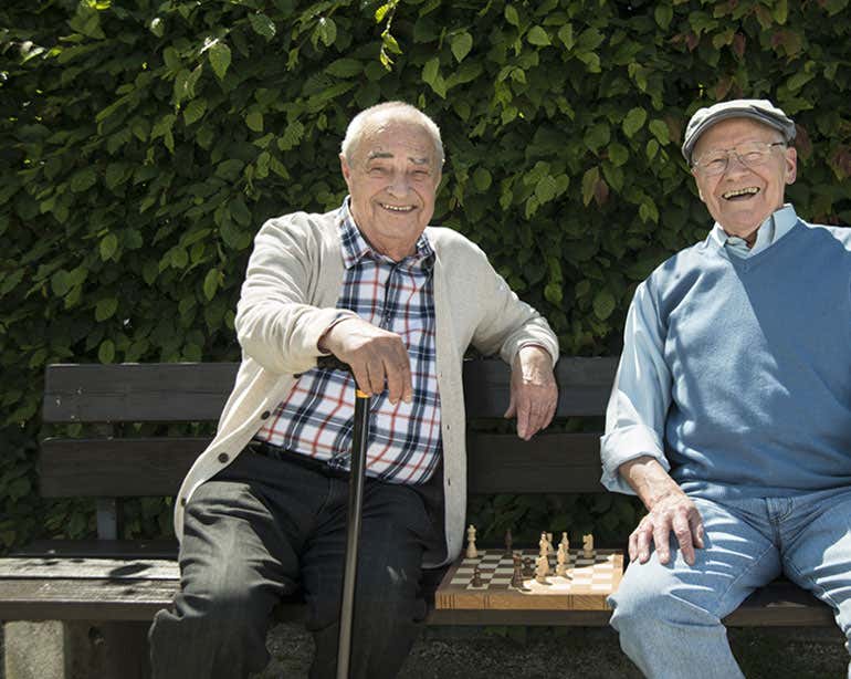 Two senior men play chess in the park, smiling at the camera. 
