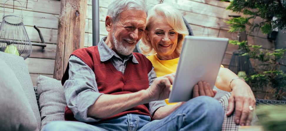 A senior couple is sitting on their patio looking at a tablet together, while smiling.