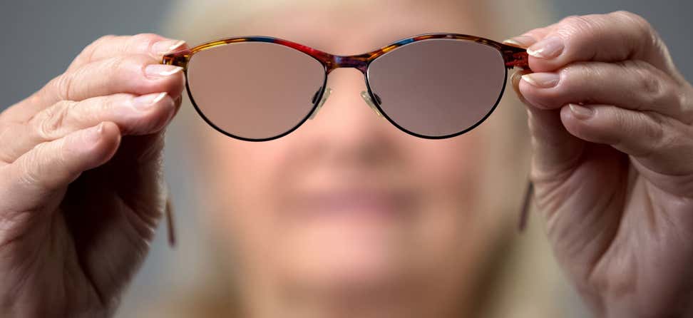 A senior Caucasian woman is holding blurred glasses up close to the camera, indicating the concept of vision loss.