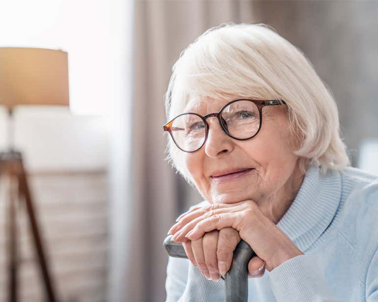 A senior Caucasian woman wearing glasses is smiling, resting her chin on her hands while holding a cane.