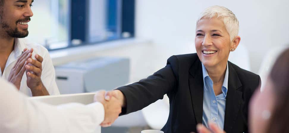 A senior business woman with short hair is smiling while she's shaking hands with another colleague.