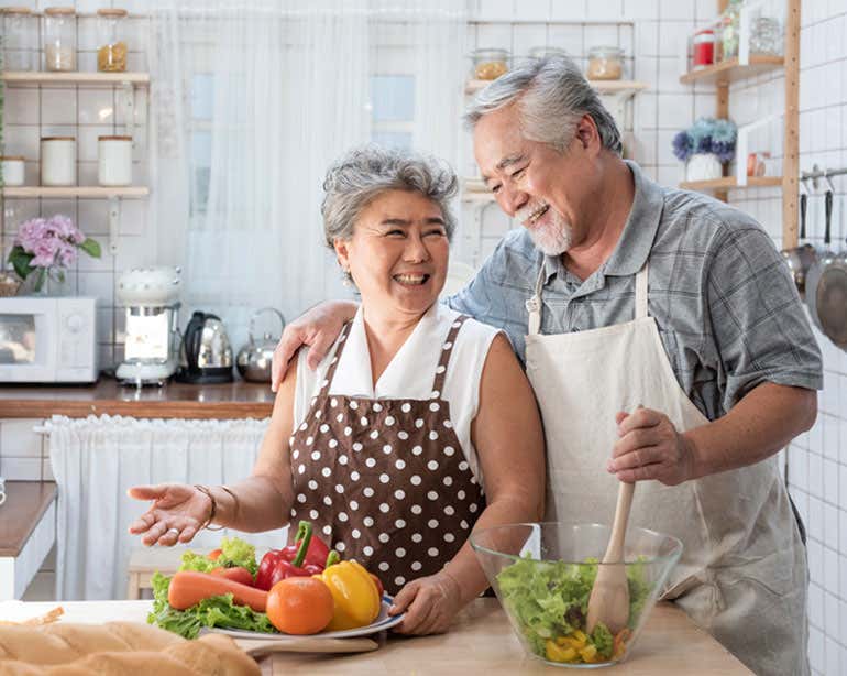 A senior Asian couple is in the kitchen smiling at each other while preparing a healthy meal.