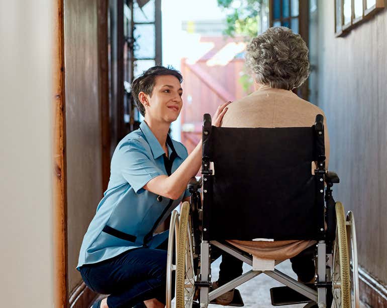 A younger caregiver with short dark hair is caring for a female senior woman in a wheelchair whose back is turned to the camera.