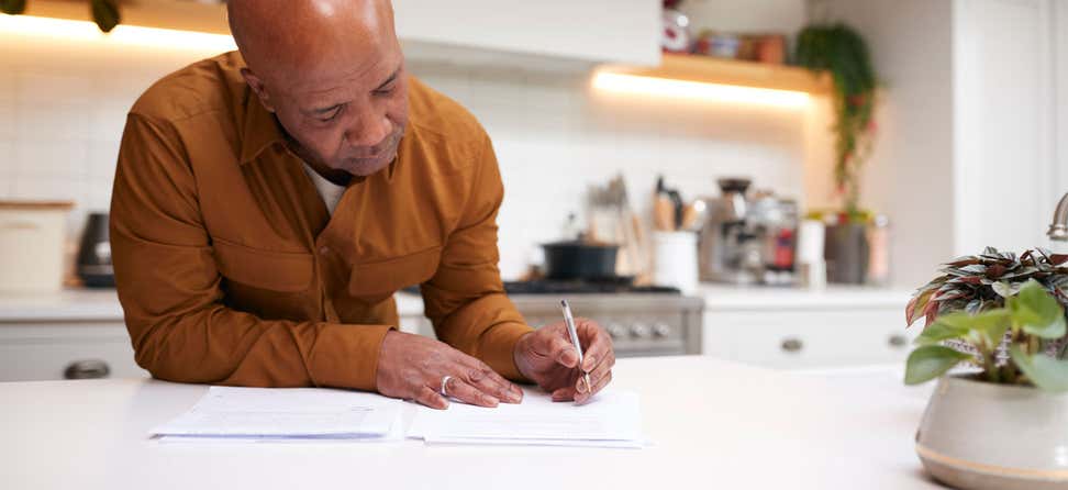 An older black man is reviewing and signing financial paperwork in his kitchen.
