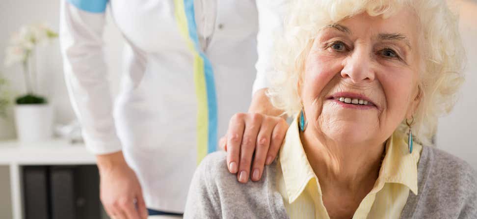 A nurse holds her hand on an older woman's shoulder, providing emotional support during her doctor's visit.