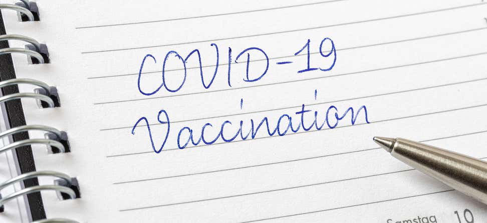 The words "COVID-19 Vaccination" written in cursive in a day planner with a pen resting on the pad.