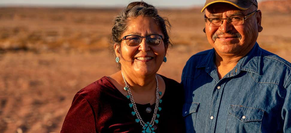 A senior Indigenous couple stands embraces each other outside, smiling.