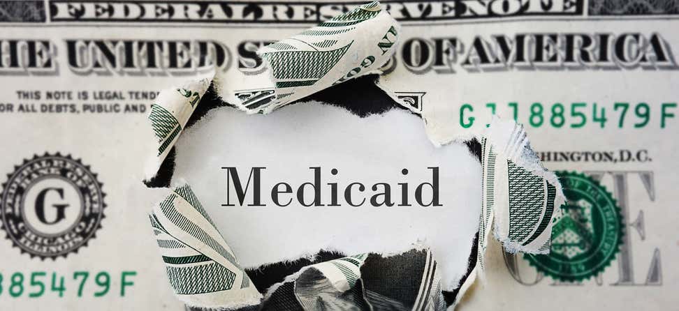 Image of a U.S. dollar bill ripped in the center with the word "Medicaid" showing through.