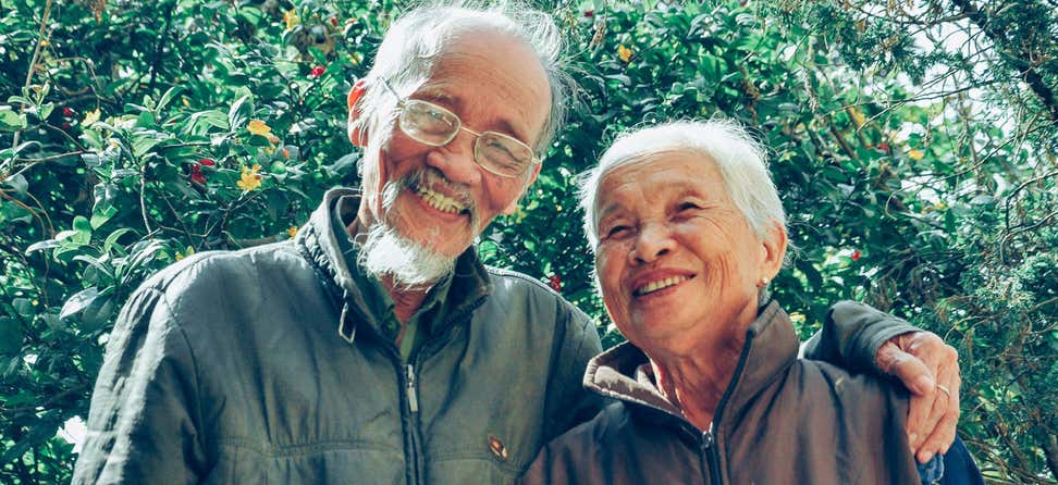 A senior Asian couple walk arm-in-arm outside, smiling and enjoying nature.