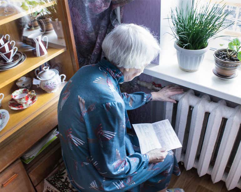 An older woman is seen looking at her utility bill while sitting next to her radiator feeling the warmth.
