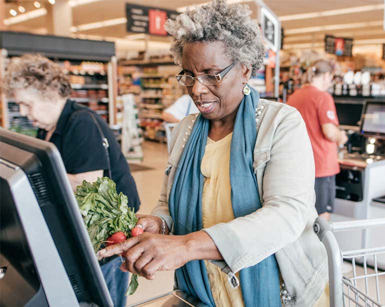 A Black senior woman is seen checking herself out at the grocery store, processing radishes over the payment kiosk. 