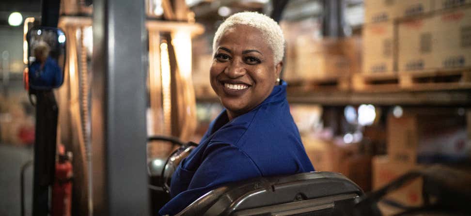 An older, black female is smiling while on the job in a production warehouse.