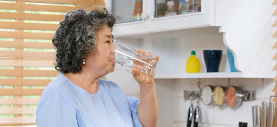 Older Asian woman drinking a glass of water in the kitchen