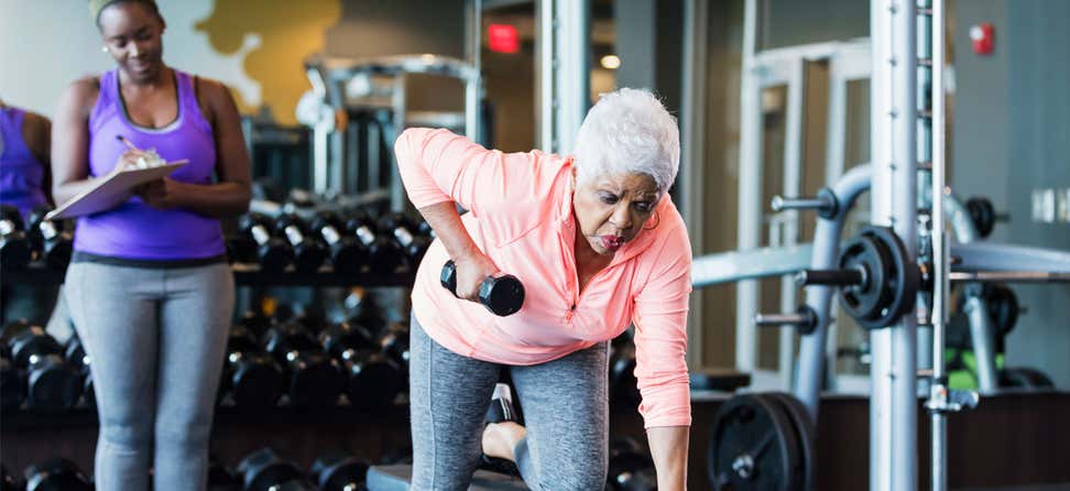 A senior Black woman is lifting weights in the gym with her trainer.