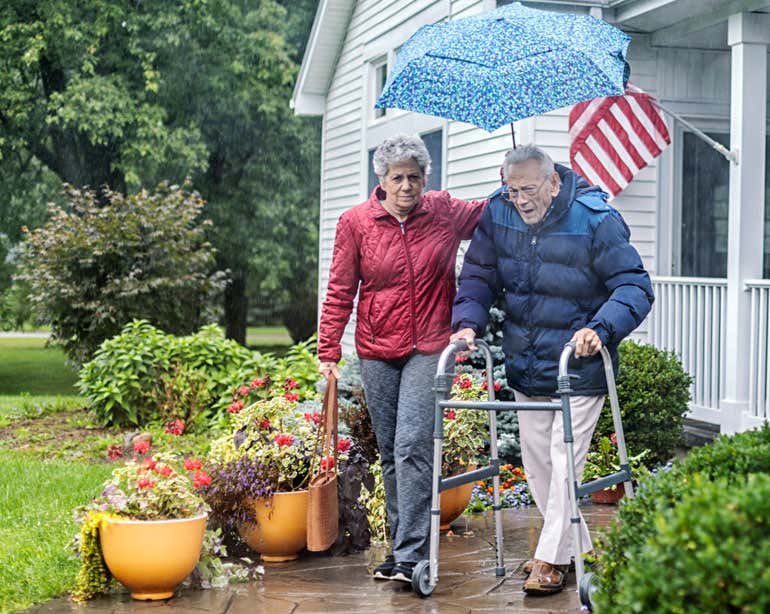 An older female caregiver helps a male veteran who's walking out of his home with a walker.