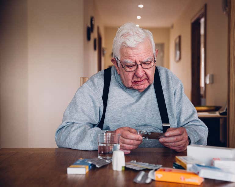 A senior man is sitting as his dining room table with all of his prescriptions.