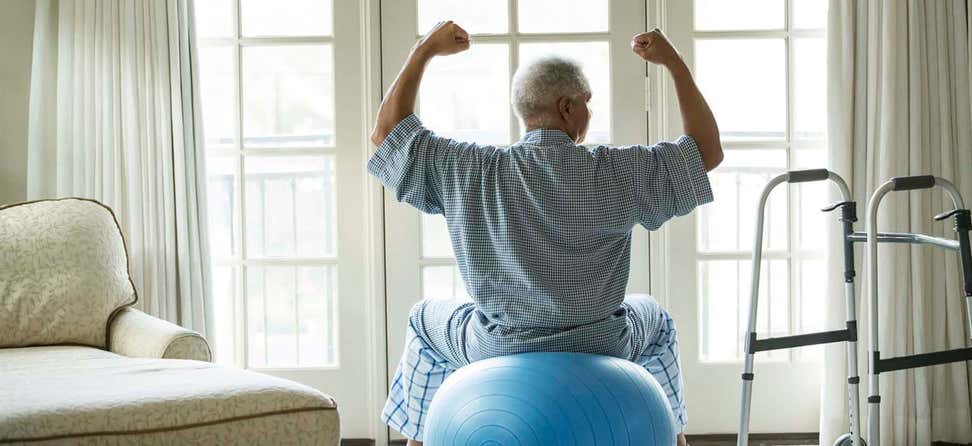 Senior African American man sits on a fitness ball at home while holding his arms up to show his strength.