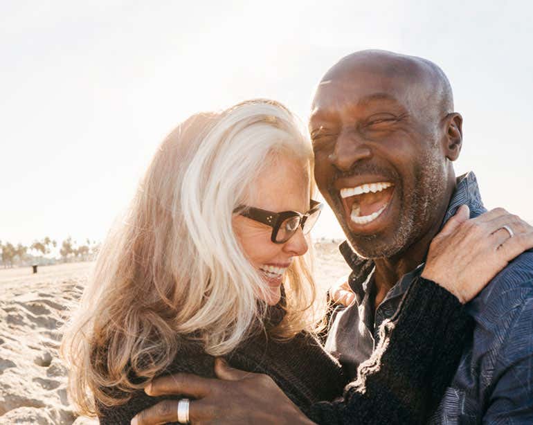 An older interracial couple is seen laughing and embracing at the beach.