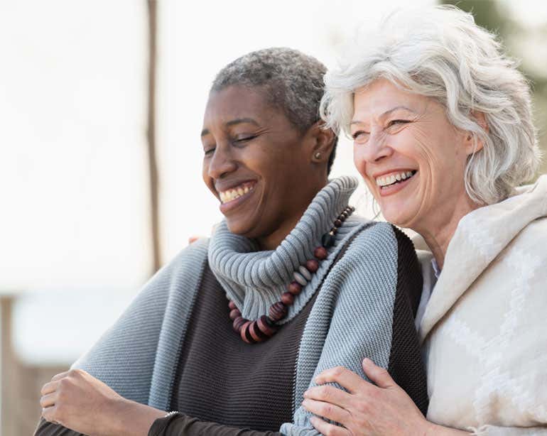 Two senior women, Black and Caucasian, are hugging each other, enjoying the day outside.
